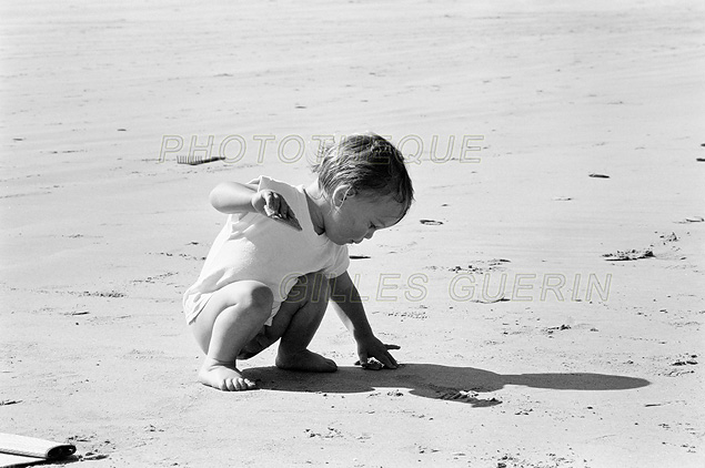 Jeune enfant accroupi pour saisir un coquillage sur une plage de sable en bord de mer - Photo noir et blanc- Presqu-île de Crozon - Bretagne - 1990